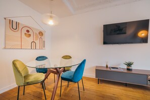 A modern dining area with a glass table, two velvet chairs, a wooden cabinet, and a flat-screen TV on the wall.