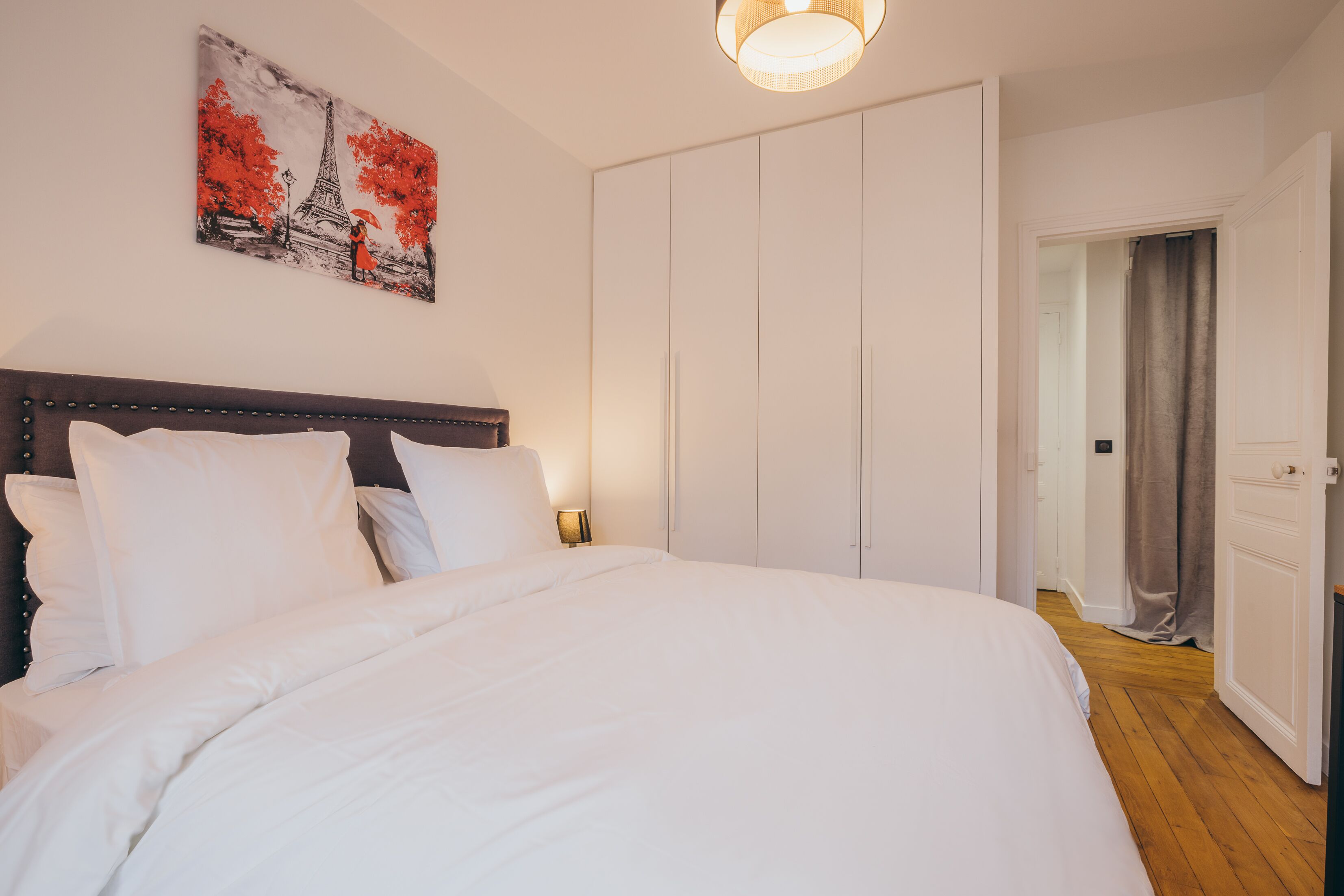 A bedroom with a large bed, white bedding, a headboard, a painting of the Eiffel Tower, and a wardrobe.