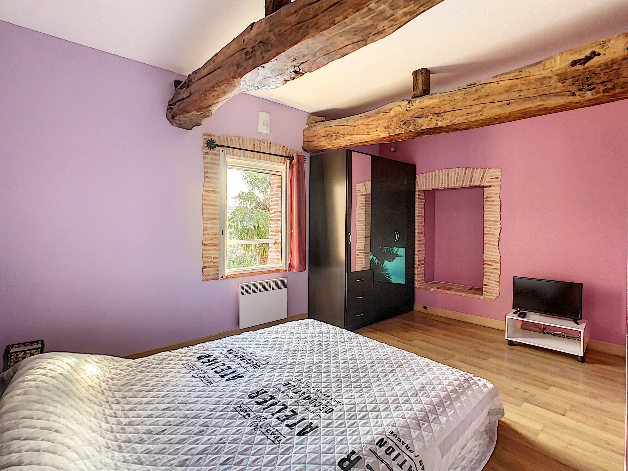 Un dormitorio con una cama, una ventana, un espejo, un televisor sobre un mueble bajo y un radiador.