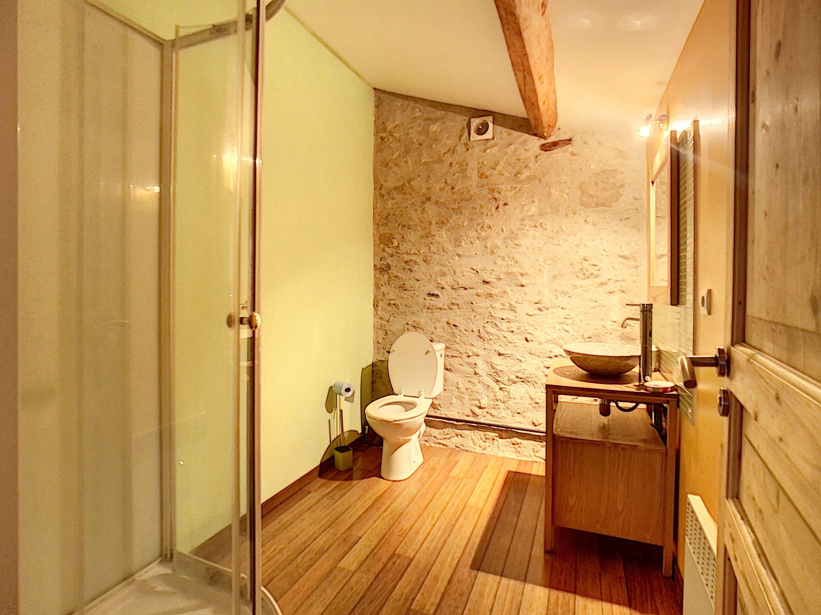 Un baño con pared de piedra, suelo de madera, cabina de ducha de vidrio, inodoro blanco y tocador de madera con lavabo.