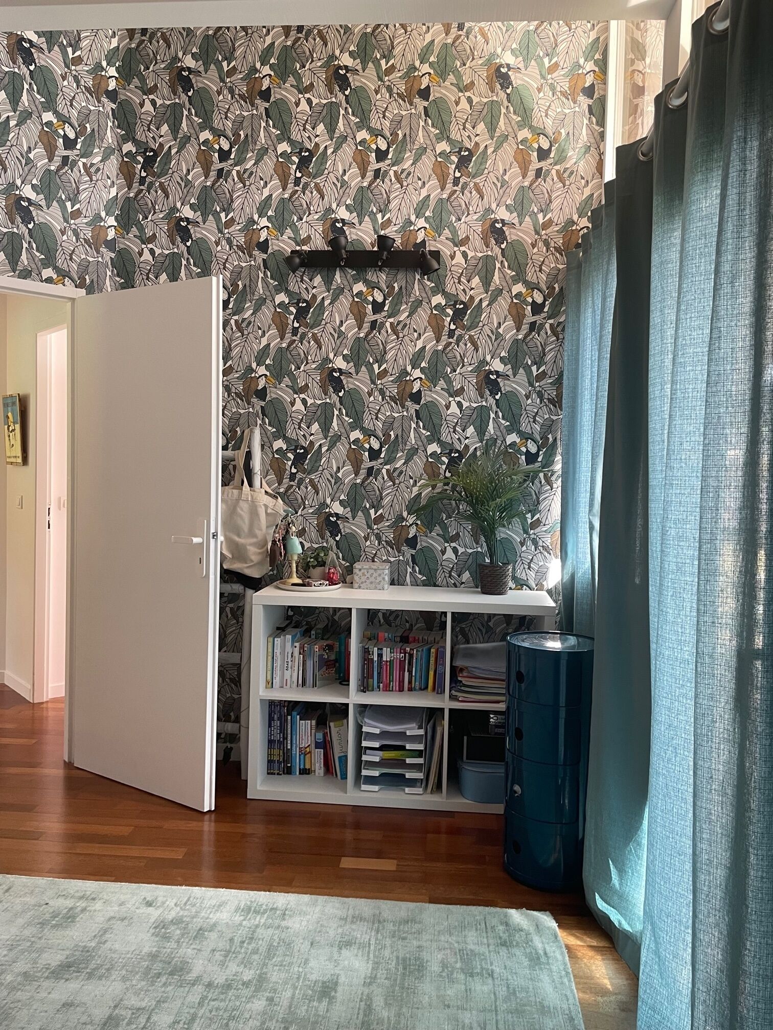 Um quarto com papel de parede estampado, uma estante branca, uma lixeira azul e uma porta entreaberta.