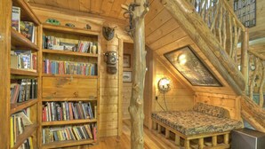 L’intérieur d’un chalet confortable, avec des murs en rondins de bois, un escalier en bois, une bibliothèque remplie de livres, un petit lit recouvert d’une couverture à motifs et une horloge fixée au mur.