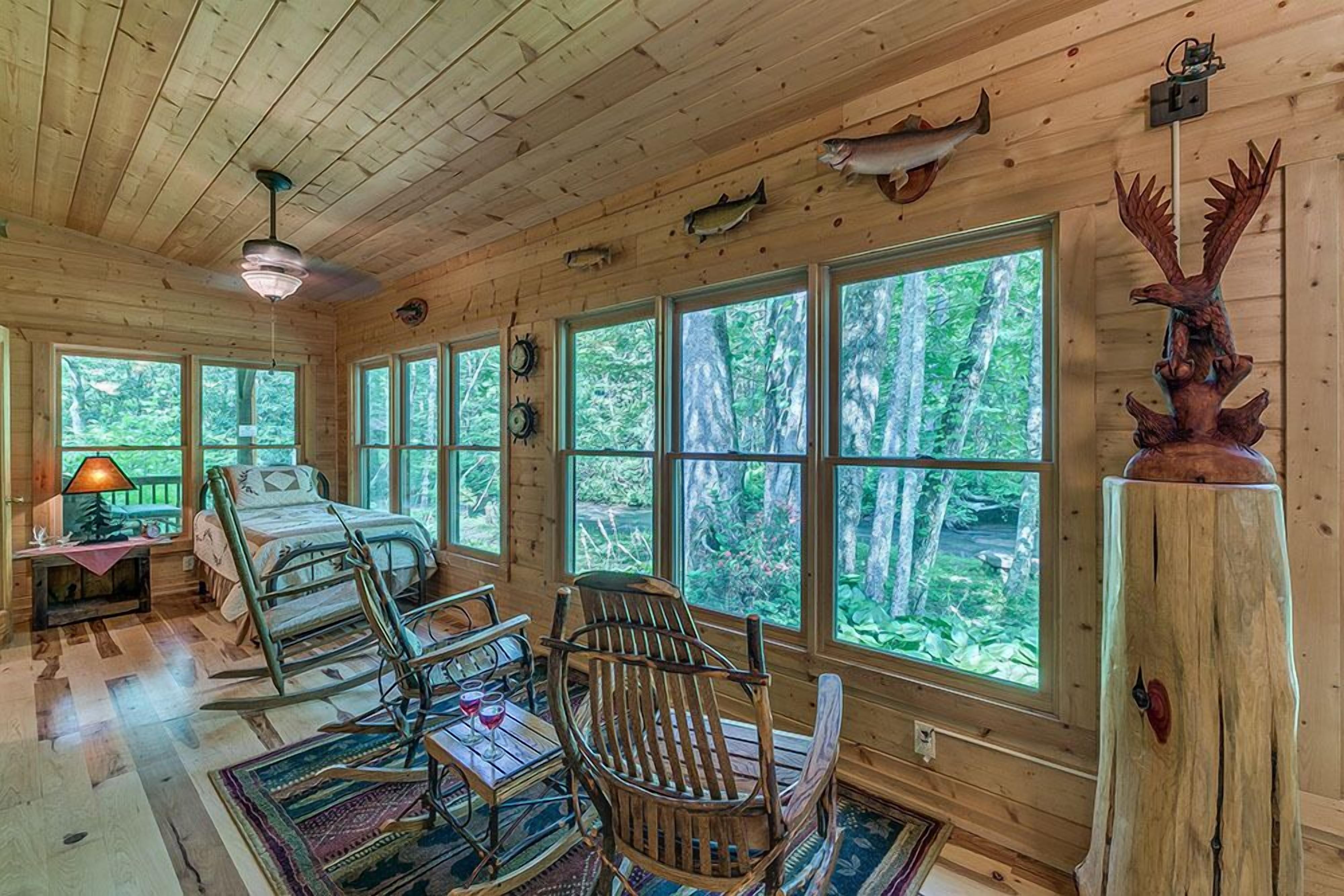 A cabin interior with a bed, rocking chairs, a table with a glass, and large windows offering a forest view.