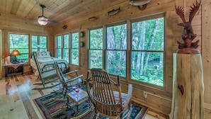 L'intérieur d'une cabane comprend un lit, des fauteuils à bascule, une table avec un verre et de grandes fenêtres donnant sur la forêt.