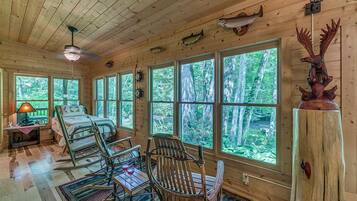 A cabin interior with a bed, rocking chairs, a table with a glass, and large windows offering a forest view.