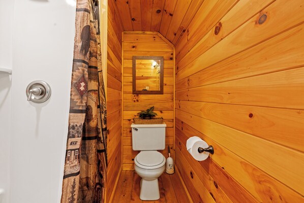 Une petite salle de bain aux murs lambrissés, équipée de toilettes, d’un panier contenant des articles de toilette et d’un miroir.