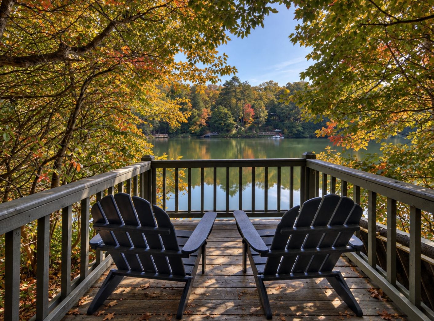 A wooden deck with two Adirondack chairs overlooking a calm lake surrounded by autumn foliage.