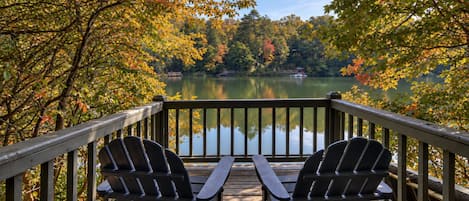 A wooden deck with two Adirondack chairs overlooking a calm lake surrounded by autumn foliage.
