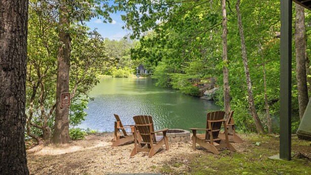 Un área de estar junto al lago con sillas Adirondack y un brasero, rodeada de árboles y vistas al lago.
