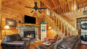 Un acogedor interior de cabaña con chimenea de piedra, muebles de cuero, televisor y escalera de madera.