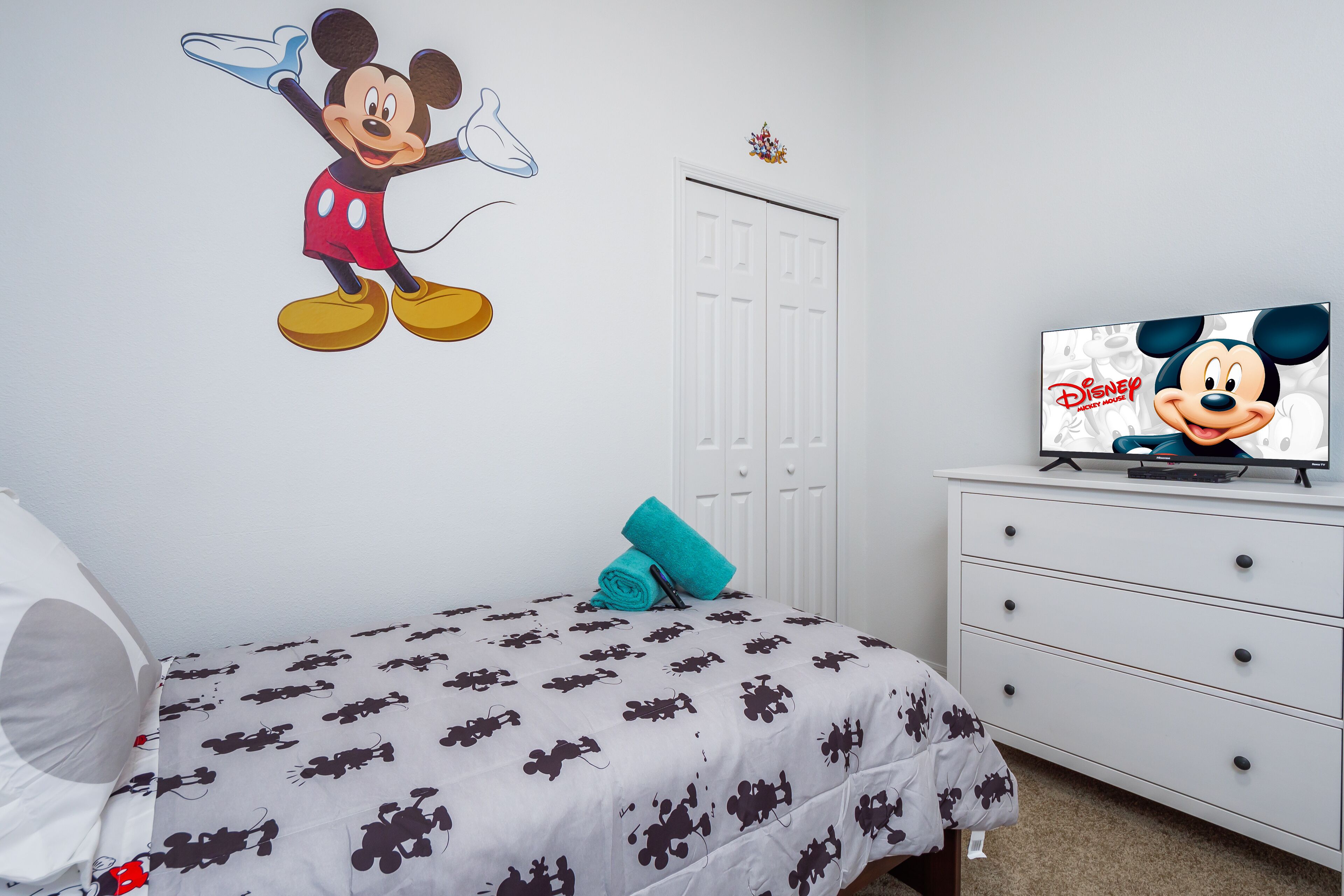 Un dormitorio con cama, televisor, cómoda blanca y un adhesivo de pared de Mickey Mouse.