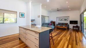 Cozinha moderna com ilha central, área de refeições com mesa de madeira, sofá cinza e TV na parede.