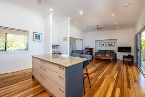 A modern kitchen with a central island, a dining area with a wooden table, a grey sofa, and a TV mounted on the wall.