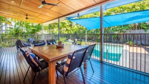Um pátio externo com mesa e cadeiras de madeira, ventoinha no teto e vista para uma piscina.