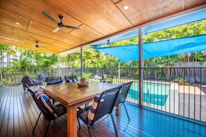 An outdoor patio with a wooden table and chairs, a ceiling fan, and a view of a swimming pool.