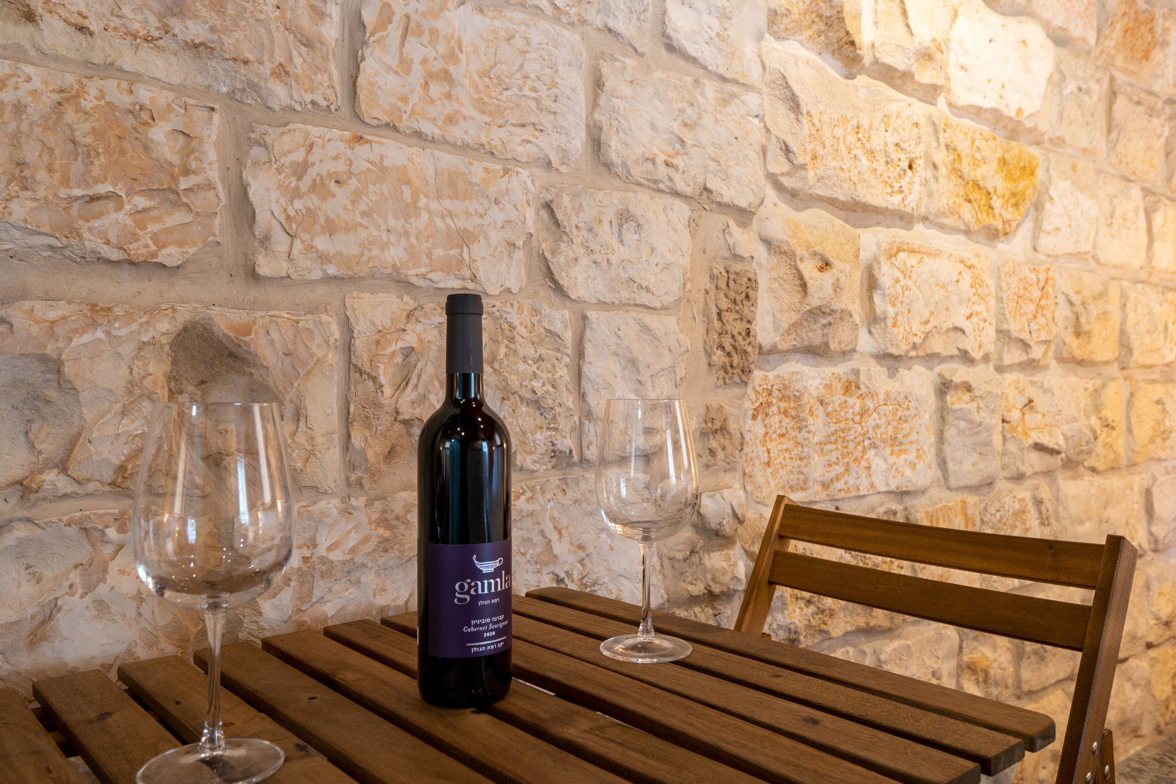 Una botella de vino y dos copas sobre una mesa de madera frente a una pared de piedra.