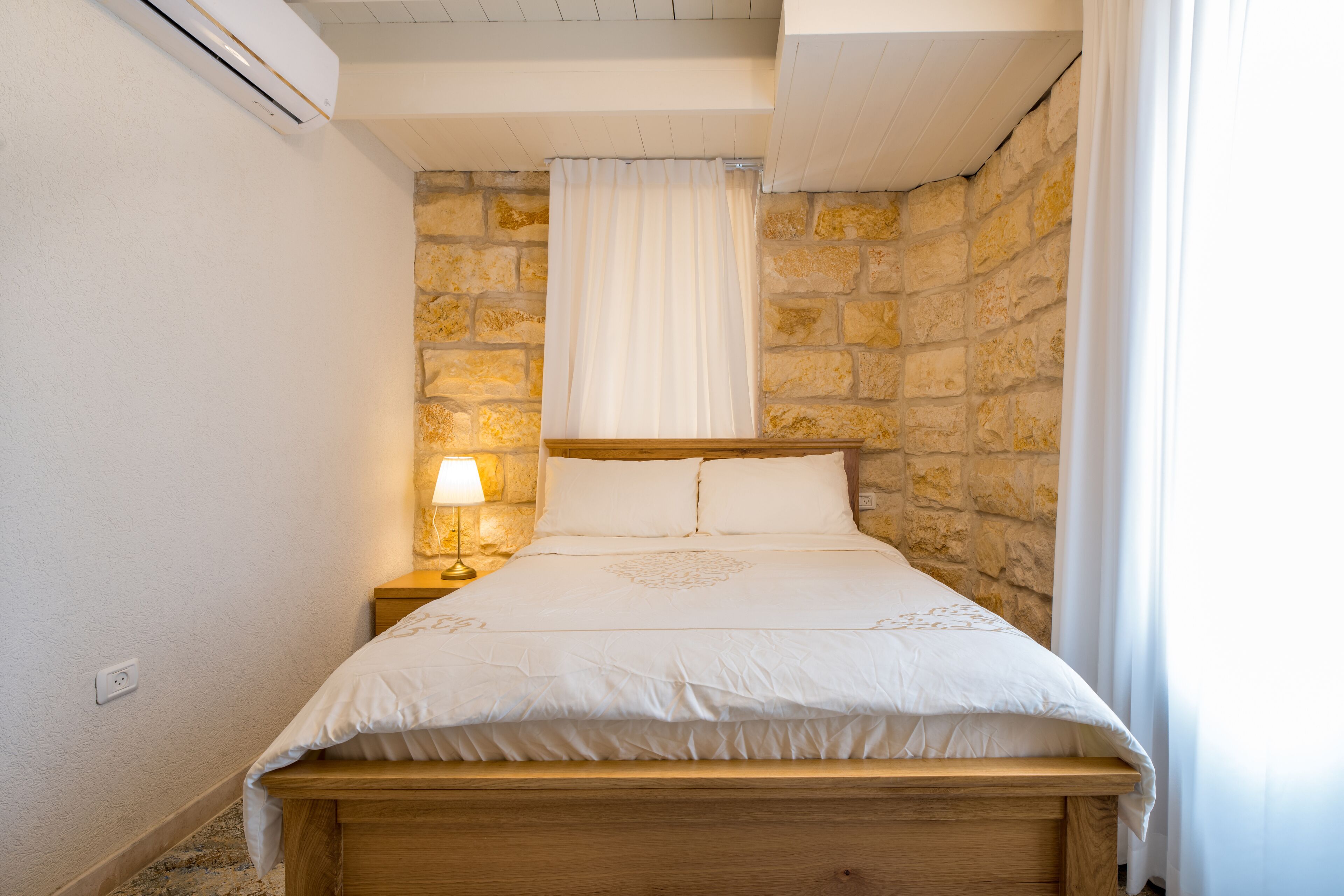 Una camera da letto con parete in pietra, struttura del letto in legno, copripiumino bianco e comodino con lampada.