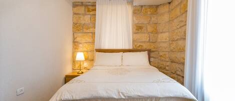 Un dormitorio con pared de piedra, estructura de cama de madera, colcha blanca y mesita de noche con lámpara.