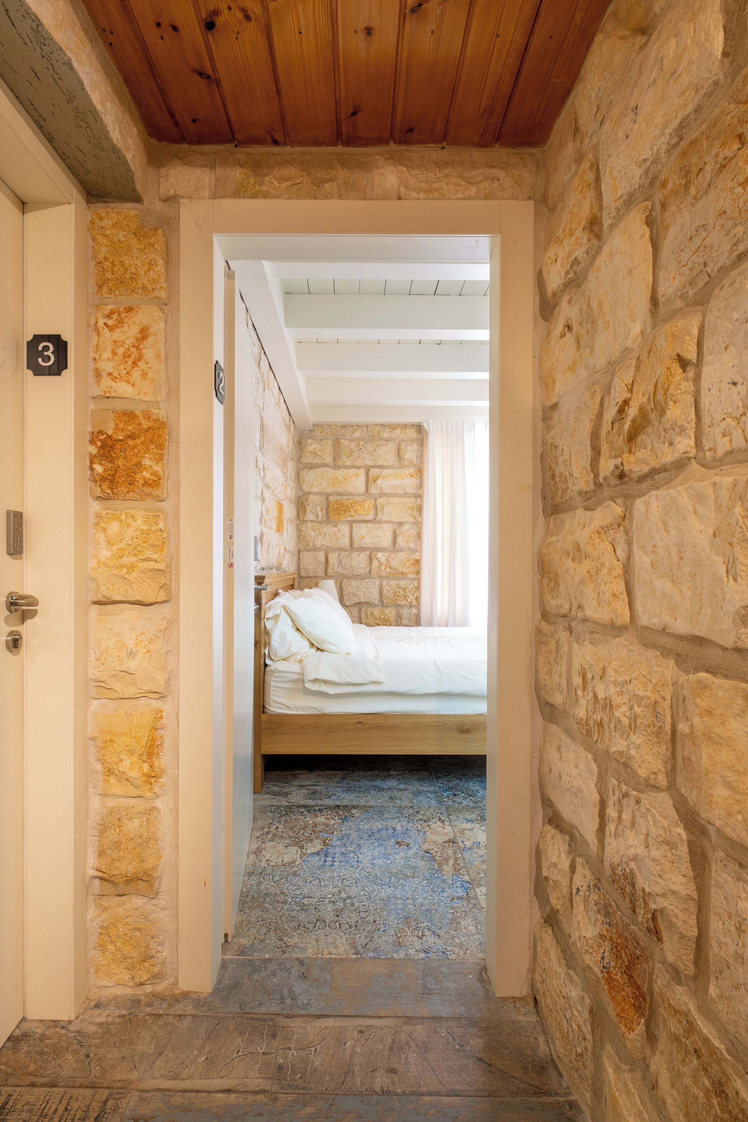 Un estrecho pasillo con pared de piedra y techo de madera, que conduce a una habitación con cama y ventana.
