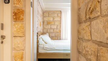 Un estrecho pasillo con pared de piedra y techo de madera, que conduce a una habitación con cama y ventana.