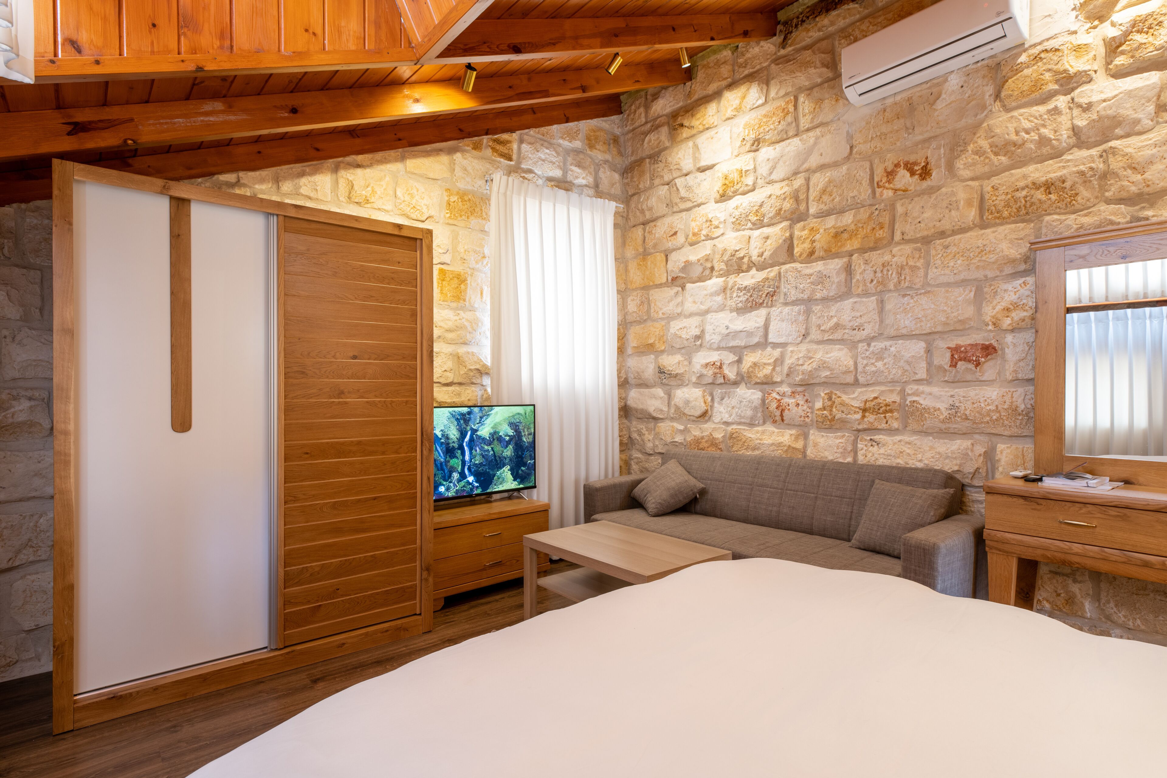 A bedroom with a wooden bed, a TV on a wooden stand, a sofa, and a stone wall.