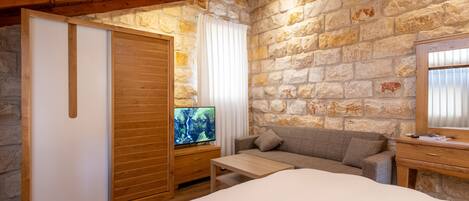 A bedroom with a wooden bed, a TV on a wooden stand, a sofa, and a stone wall.