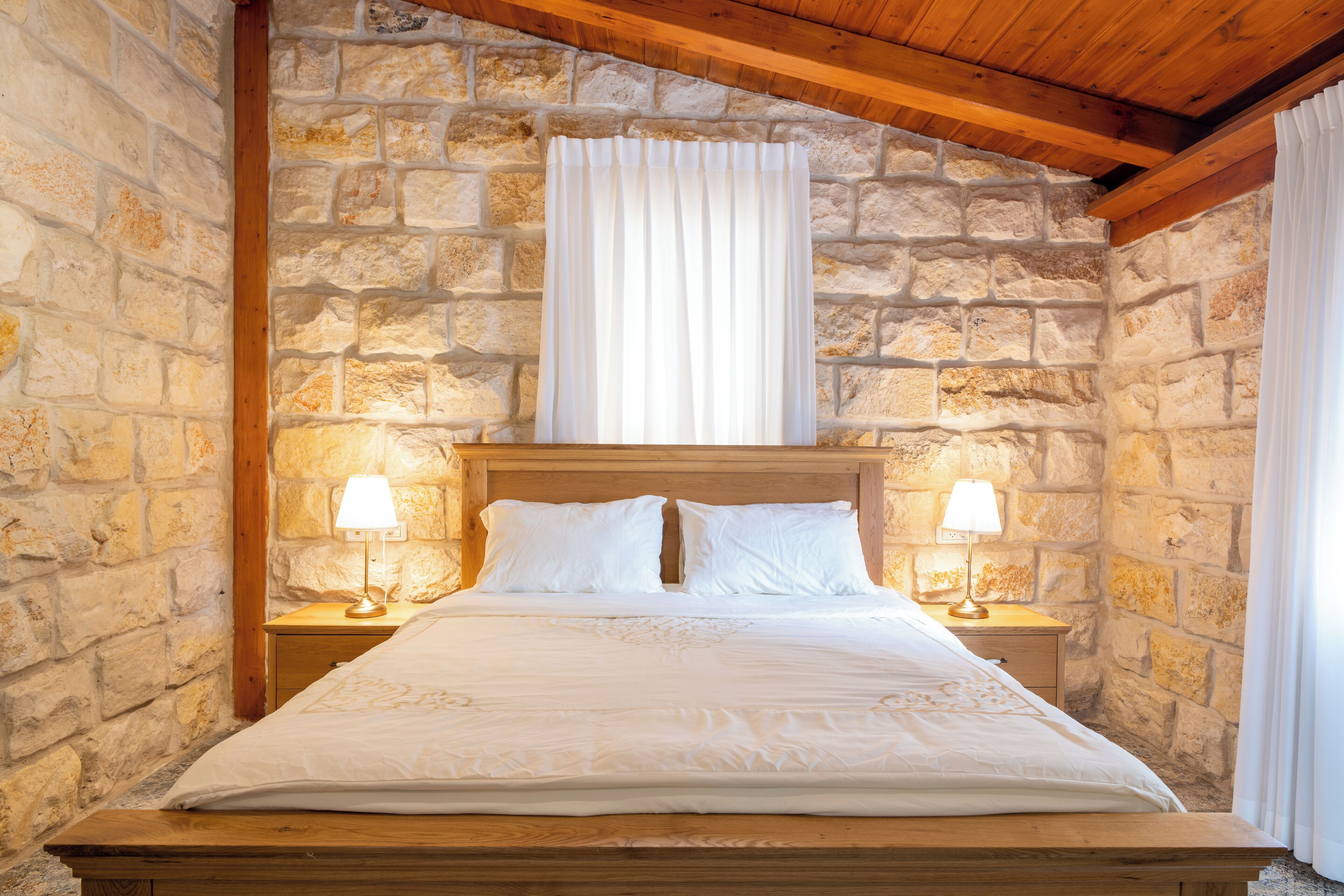 A bed with white linens and two bedside lamps, set against a stone wall with a wooden ceiling.