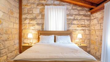 A bed with white linens and two bedside lamps, set against a stone wall with a wooden ceiling.