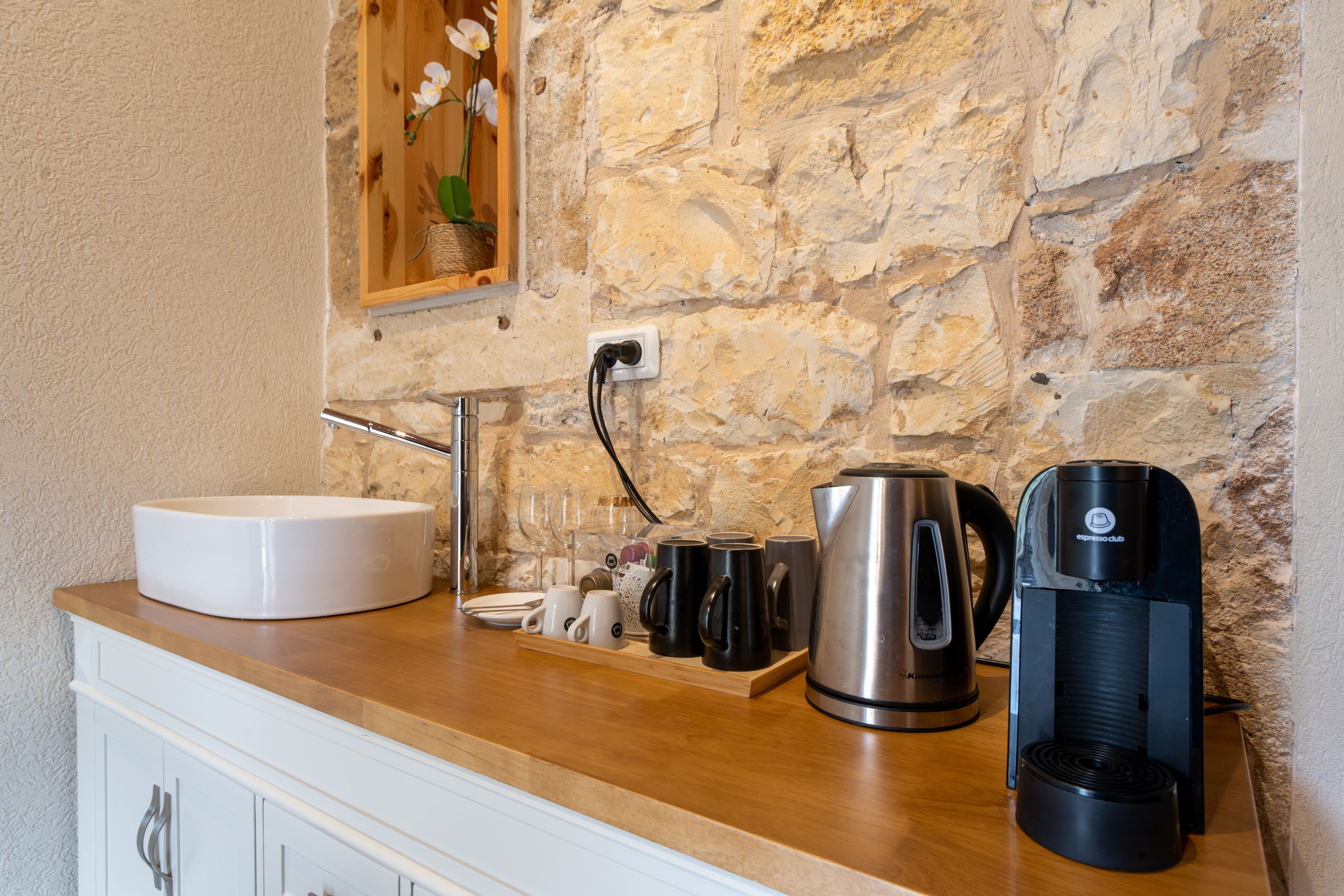 Una cocina con pared de piedra, un lavamanos blanco, una cafetera y un hervidor.
