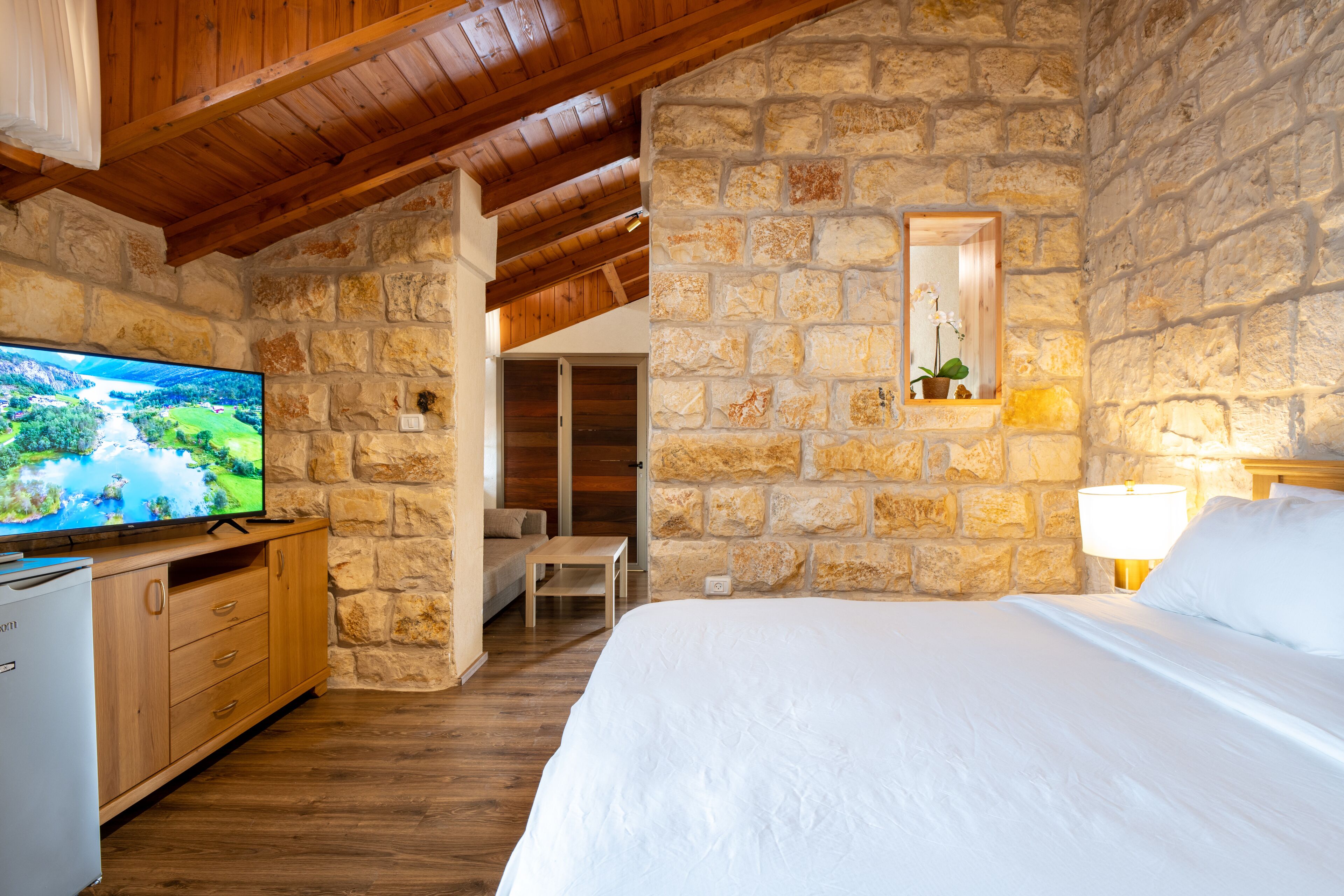 A bedroom with stone walls, a wooden ceiling, a bed, a TV, a desk, and a lamp.