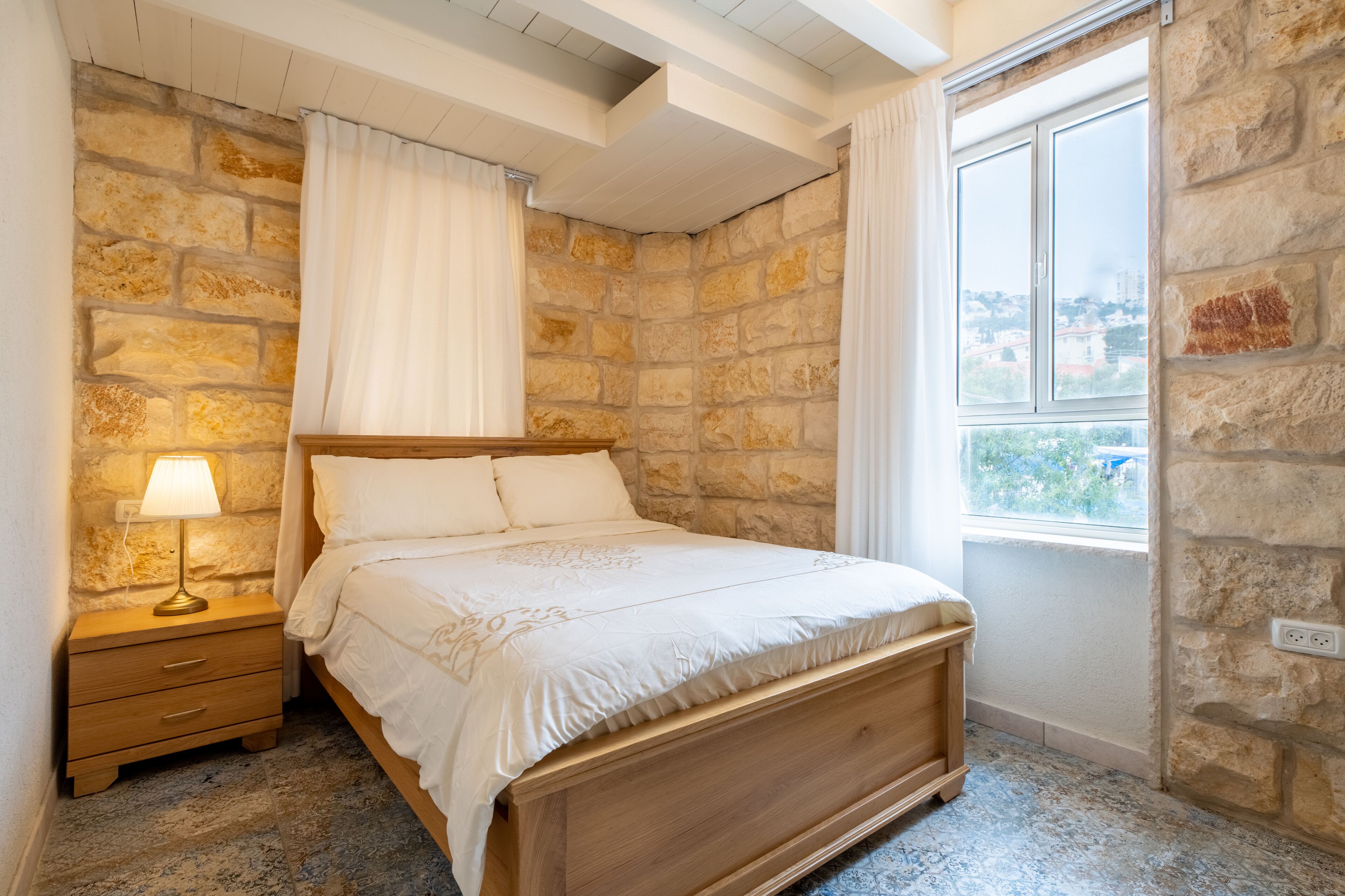 A bedroom with a stone wall, a wooden bed, a nightstand with a lamp, and a window with curtains.