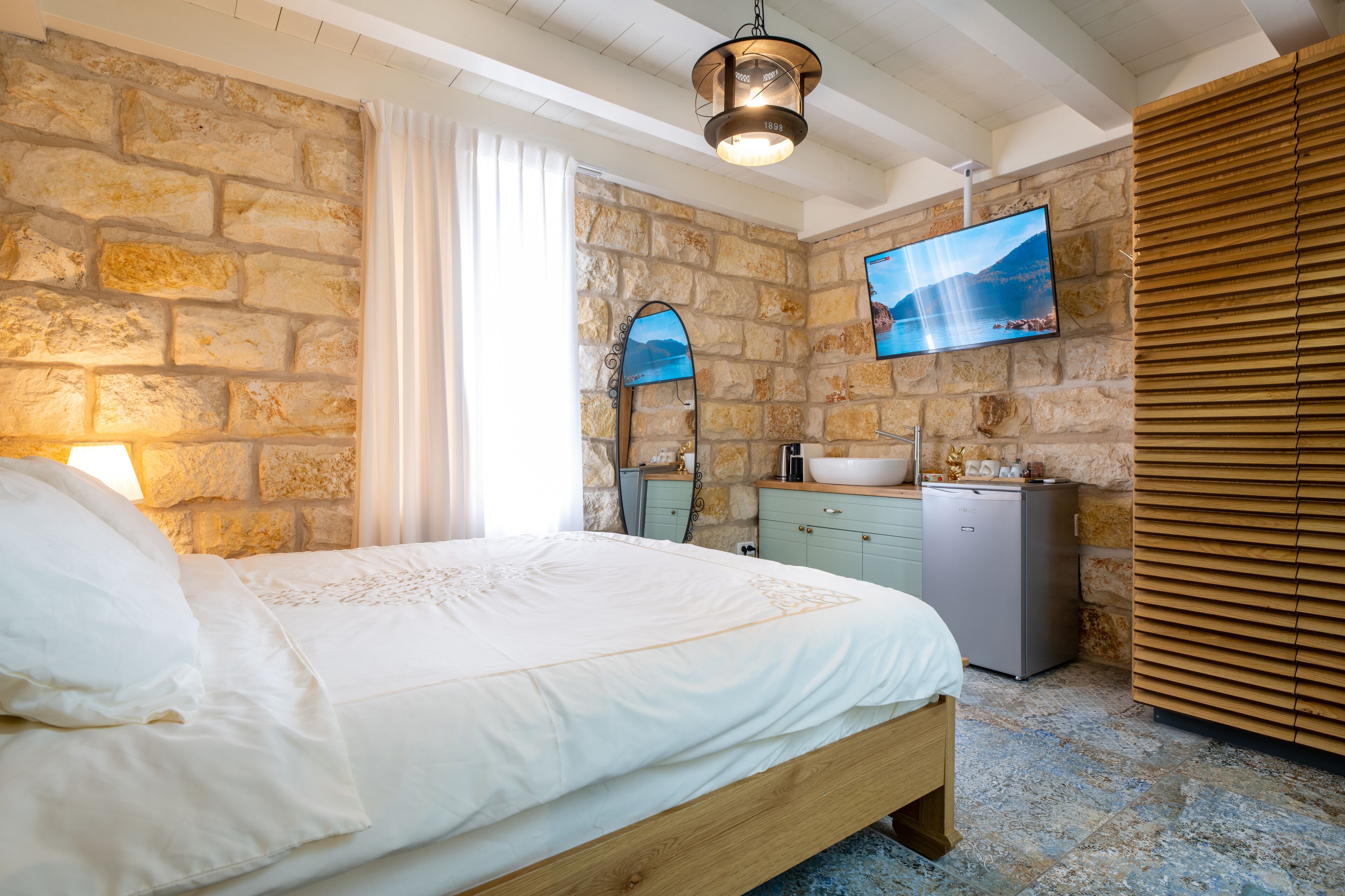 Un dormitorio con pared de piedra, cama con ropa blanca, cabecero de madera, lavamanos, espejo, televisor y pared de listones de madera.