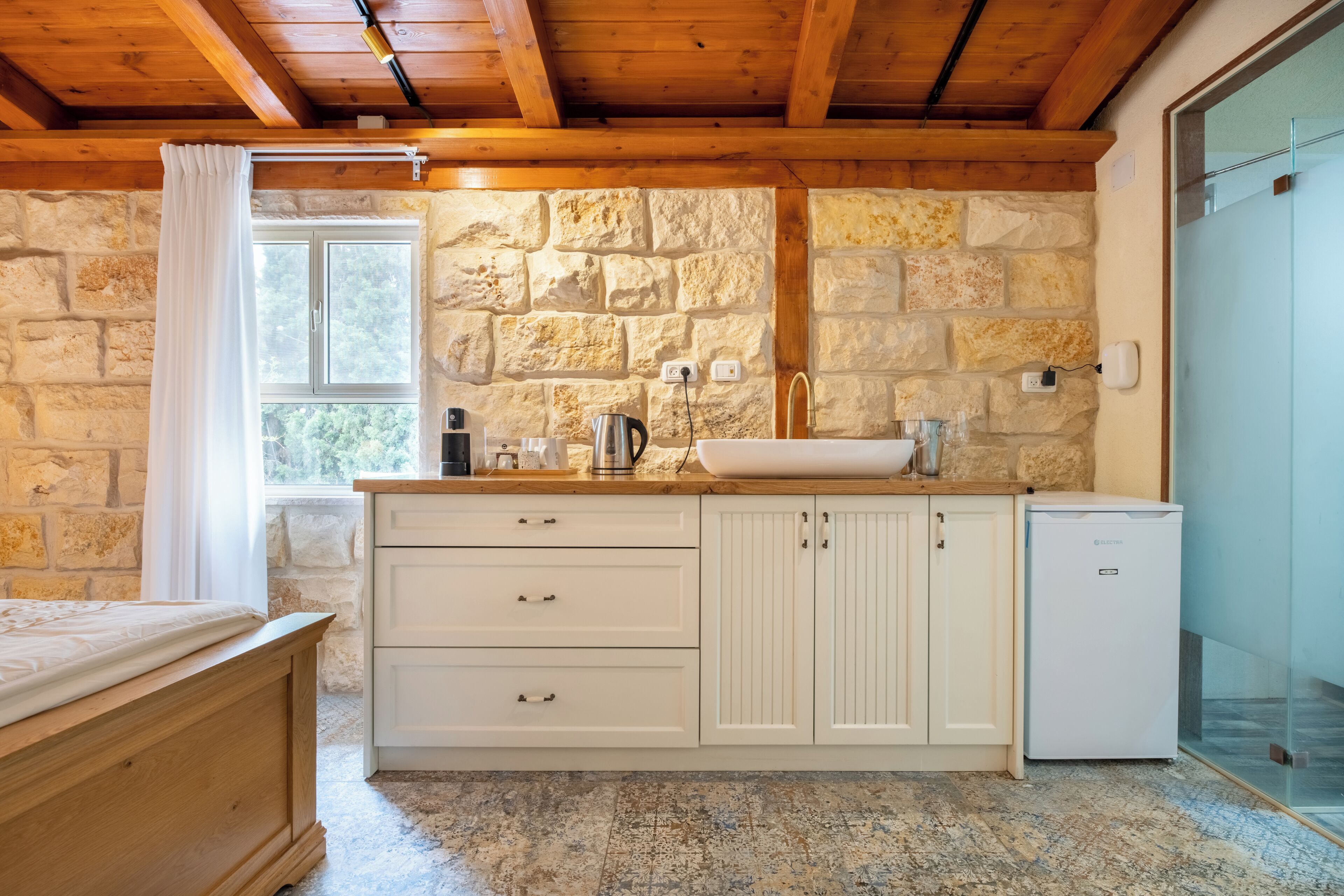 A rustic kitchen with stone walls, a wooden ceiling, a sink, and a small refrigerator.