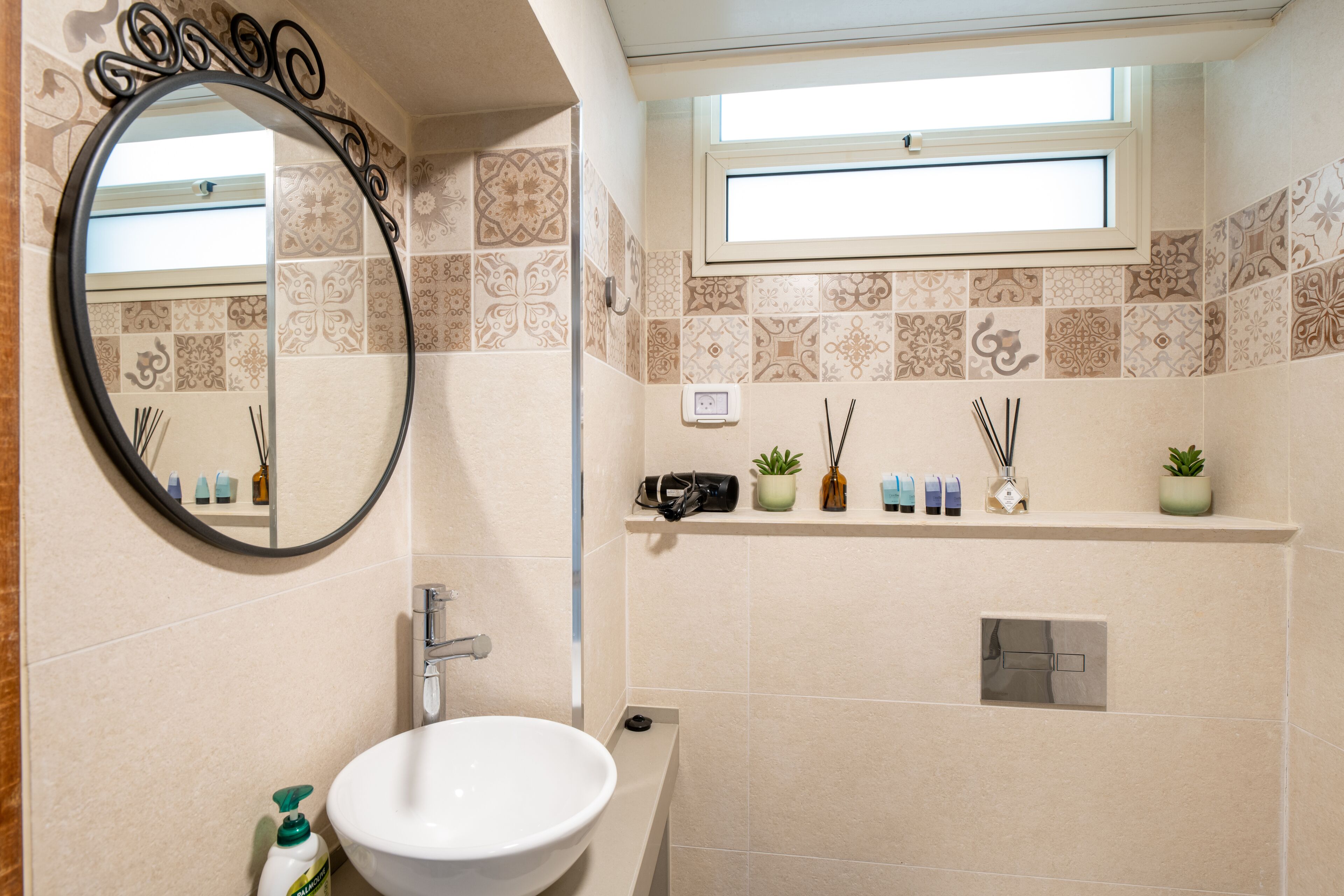 A bathroom with a round mirror, a sink, a window, and a shelf with toiletries.
