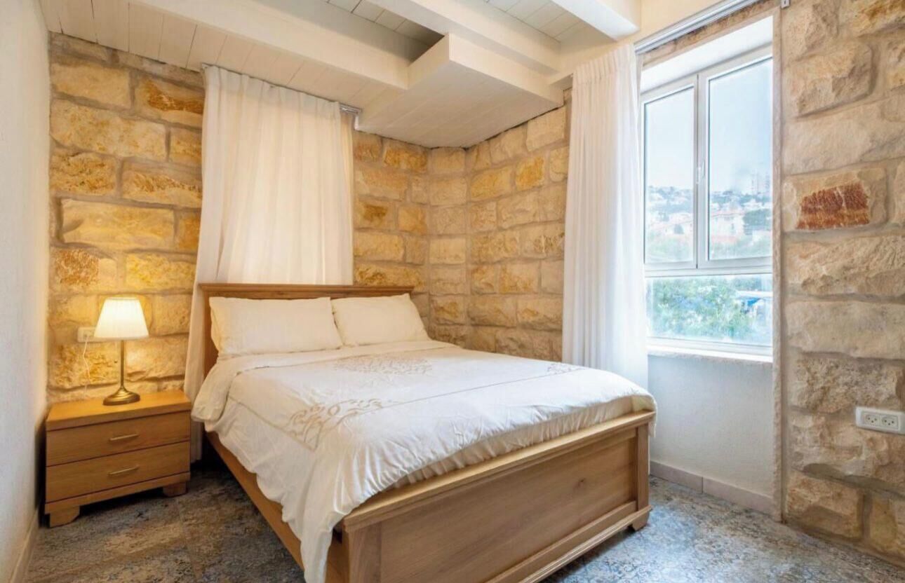 Un dormitorio con pared de piedra, una cama de madera, una mesita de noche con una lámpara y una ventana con vistas.