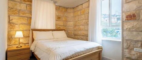 A bedroom with a stone wall, a wooden bed, a nightstand with a lamp, and a window with a view.