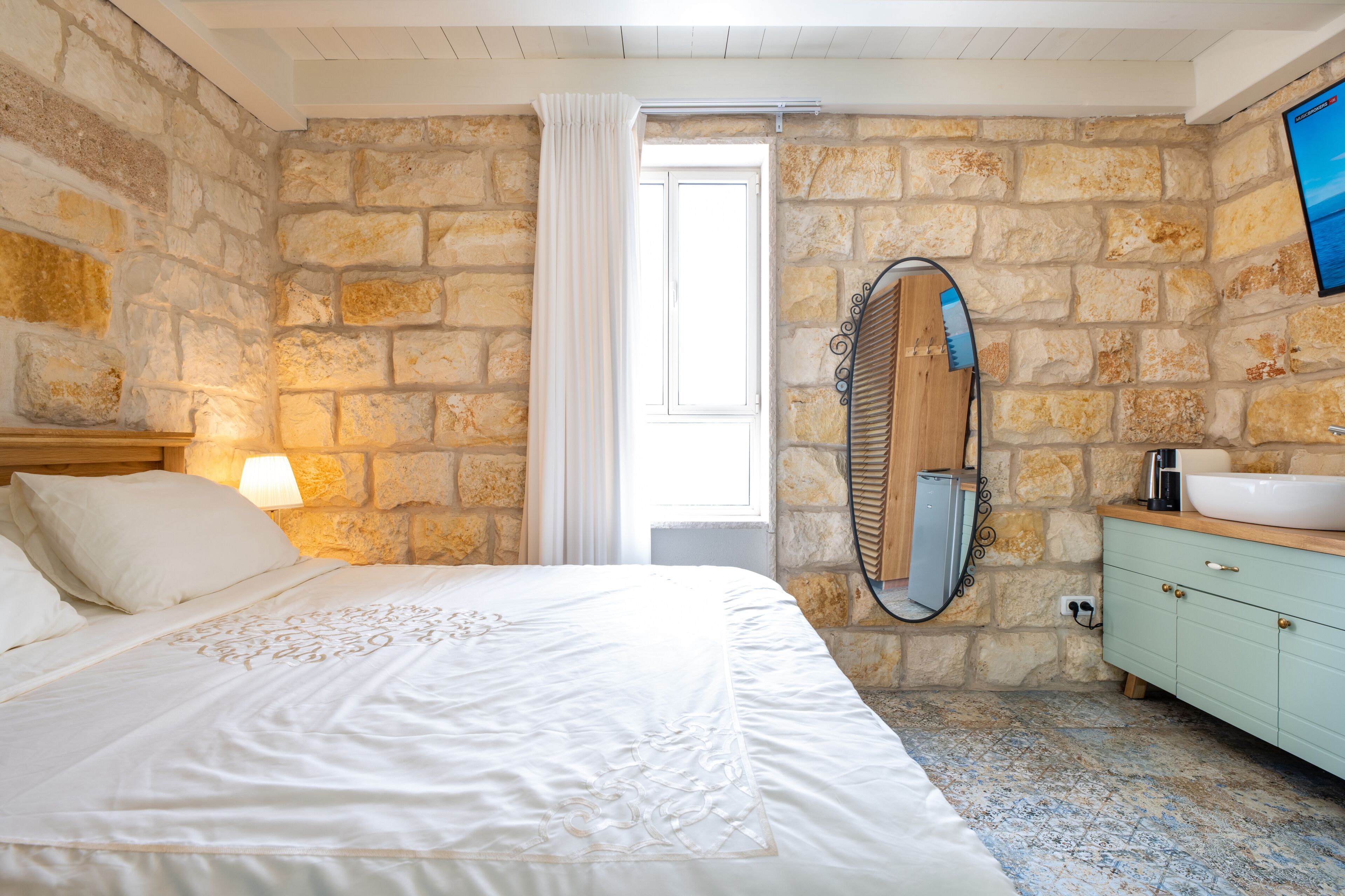 A bedroom with a stone wall, a bed with white bedding, a wooden headboard, a nightstand with a lamp, a bathroom with a sink, a mirror, and a tiled floor.