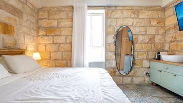 A bedroom with a stone wall, a bed with white bedding, a wooden headboard, a nightstand with a lamp, a bathroom with a sink, a mirror, and a tiled floor.