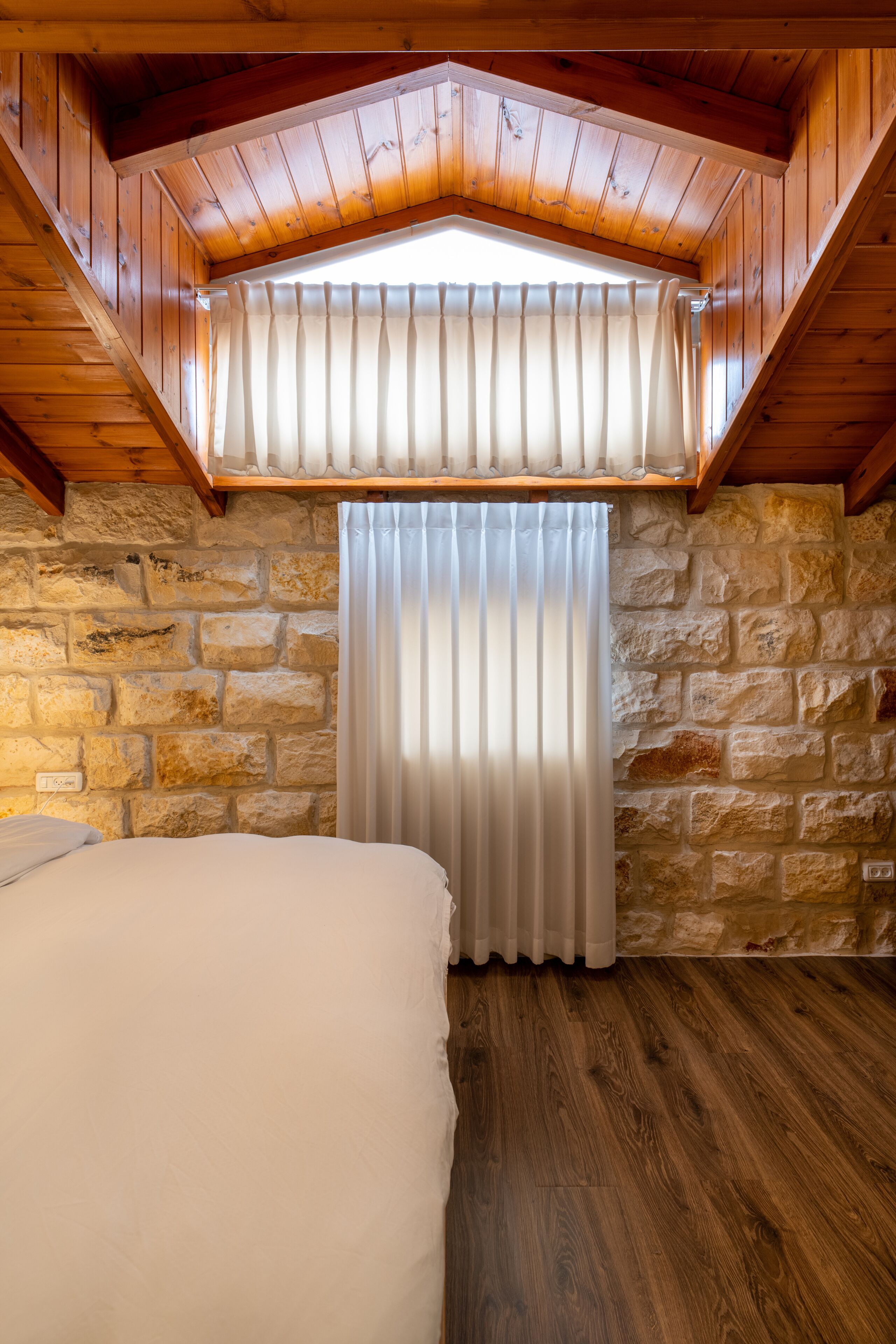 A bedroom with a stone wall, wooden ceiling, and a window with curtains.