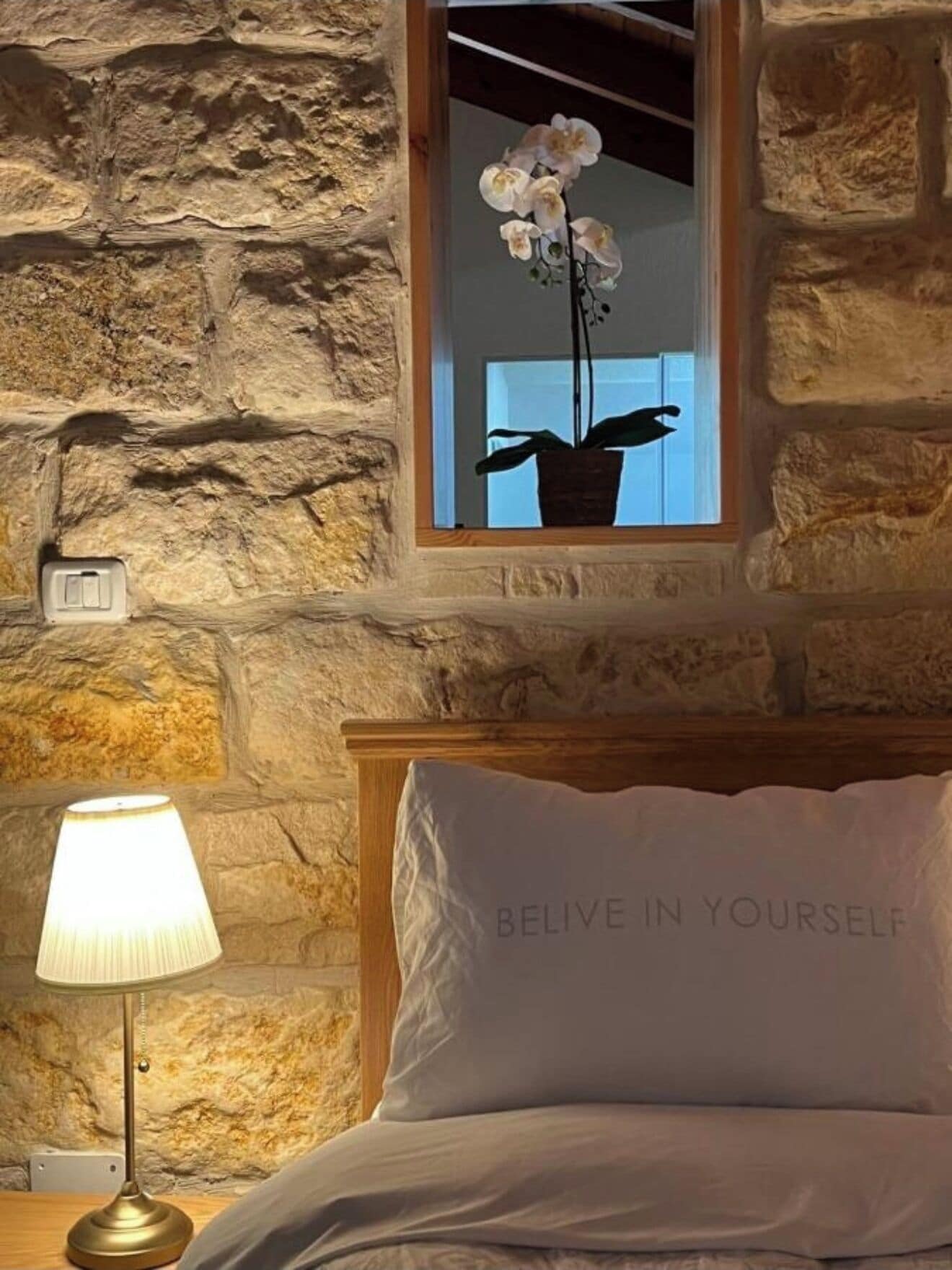 A bedroom with a stone wall, a window with an orchid plant, a bedside lamp, and a pillow with the text "BELIEVE IN YOURSELF".