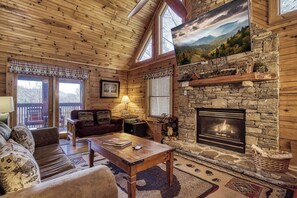 A log cabin interior with a stone fireplace, wooden ceiling, and a large window overlooking a mountainous landscape.