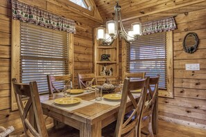 A wooden dining room with a table set for four, flanked by two windows with blinds, a chandelier, and a shelf with decorative items.