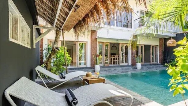 Zona junto a la piscina con sillones de descanso, techo de paja y vista a un edificio con amplias ventanas.