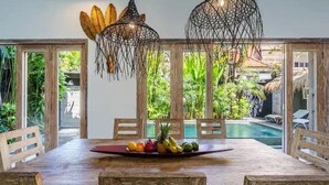 Una mesa de comedor de madera con una bandeja de frutas, rodeada de sillas de madera, en una habitación con ventanas grandes y lámparas colgantes de fibra.