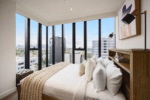 Un dormitorio con una cama grande, una mesita de noche de madera, una mesilla auxiliar y vistas de la ciudad a través de ventanales.