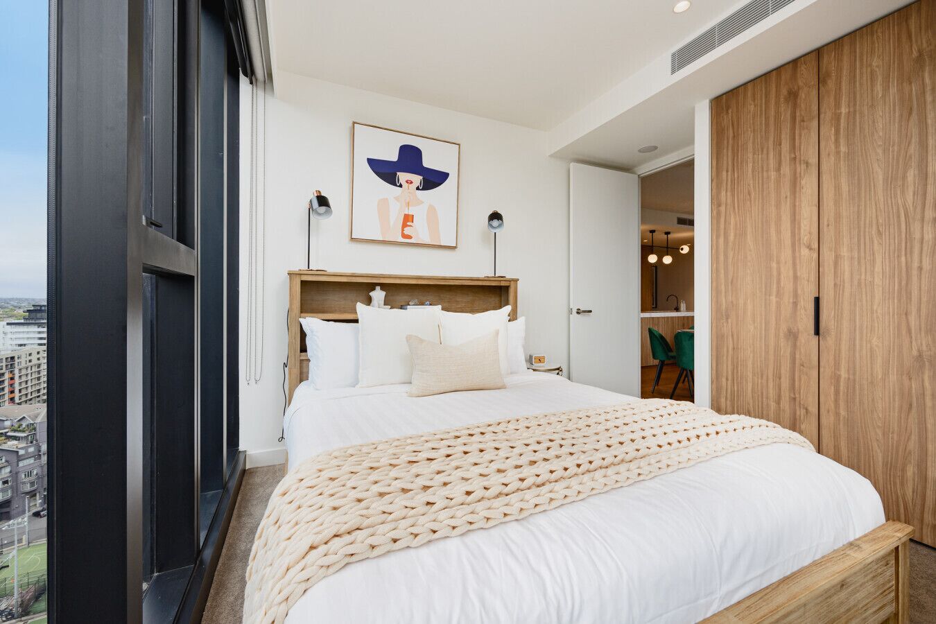 A modern bedroom with a large bed, a wooden headboard, a city view, and a wooden wardrobe.