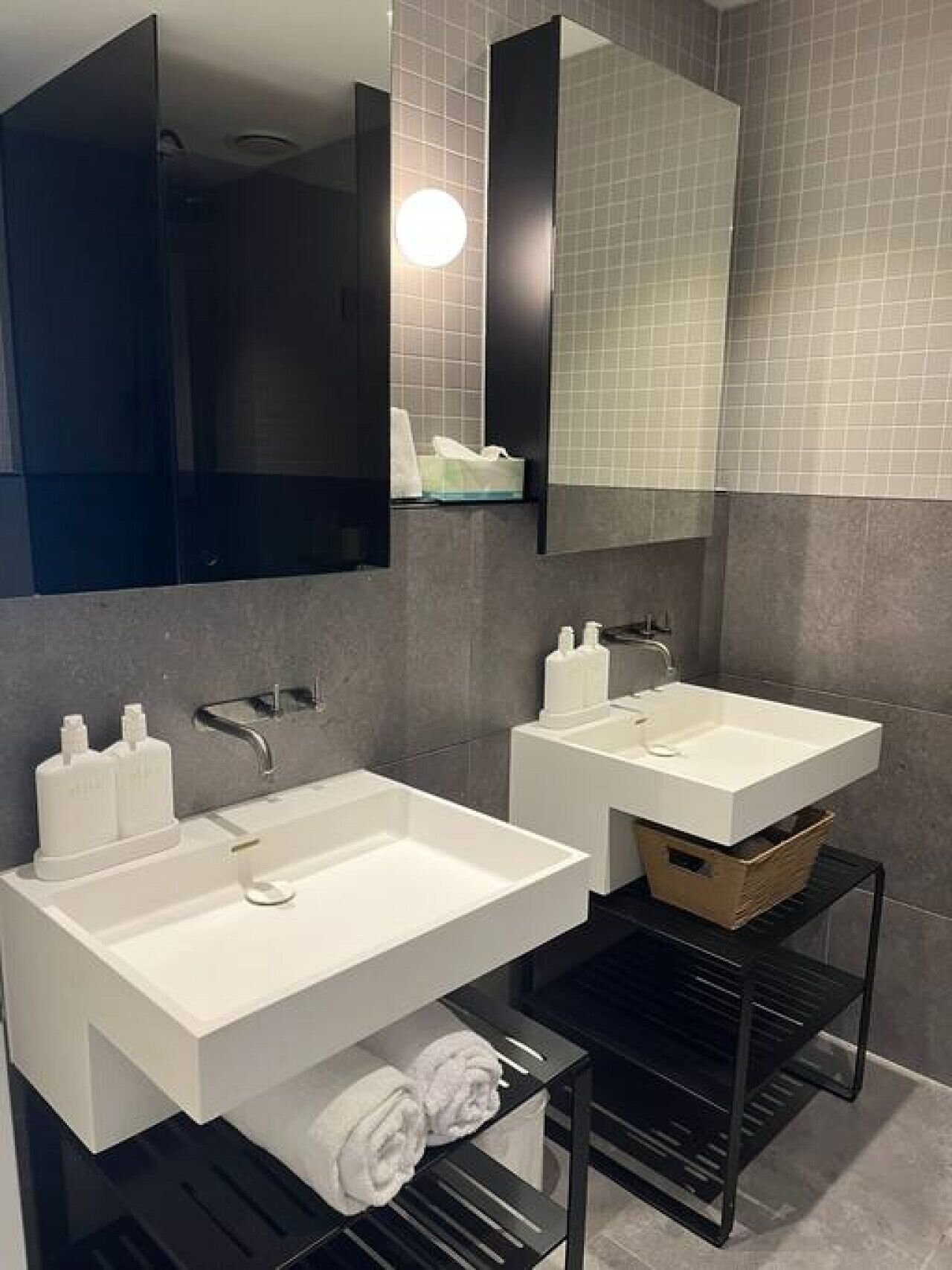 Une salle de bain moderne avec deux lavabos blancs, un grand miroir et une étagère noire où sont posées des serviettes.