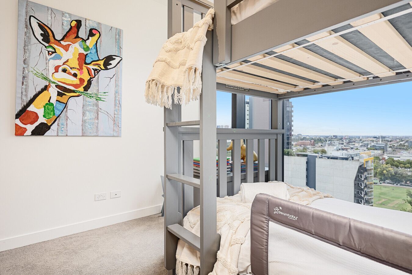 Un balcon donnant sur un paysage urbain, un tableau coloré représentant une girafe et un lit superposé avec un matelas de la marque « Dreambaby ».