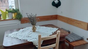 Un comedor con mesa de madera, banco y sillas. Hay plantas en macetas en el alféizar de la ventana y un florero con flores sobre la mesa.