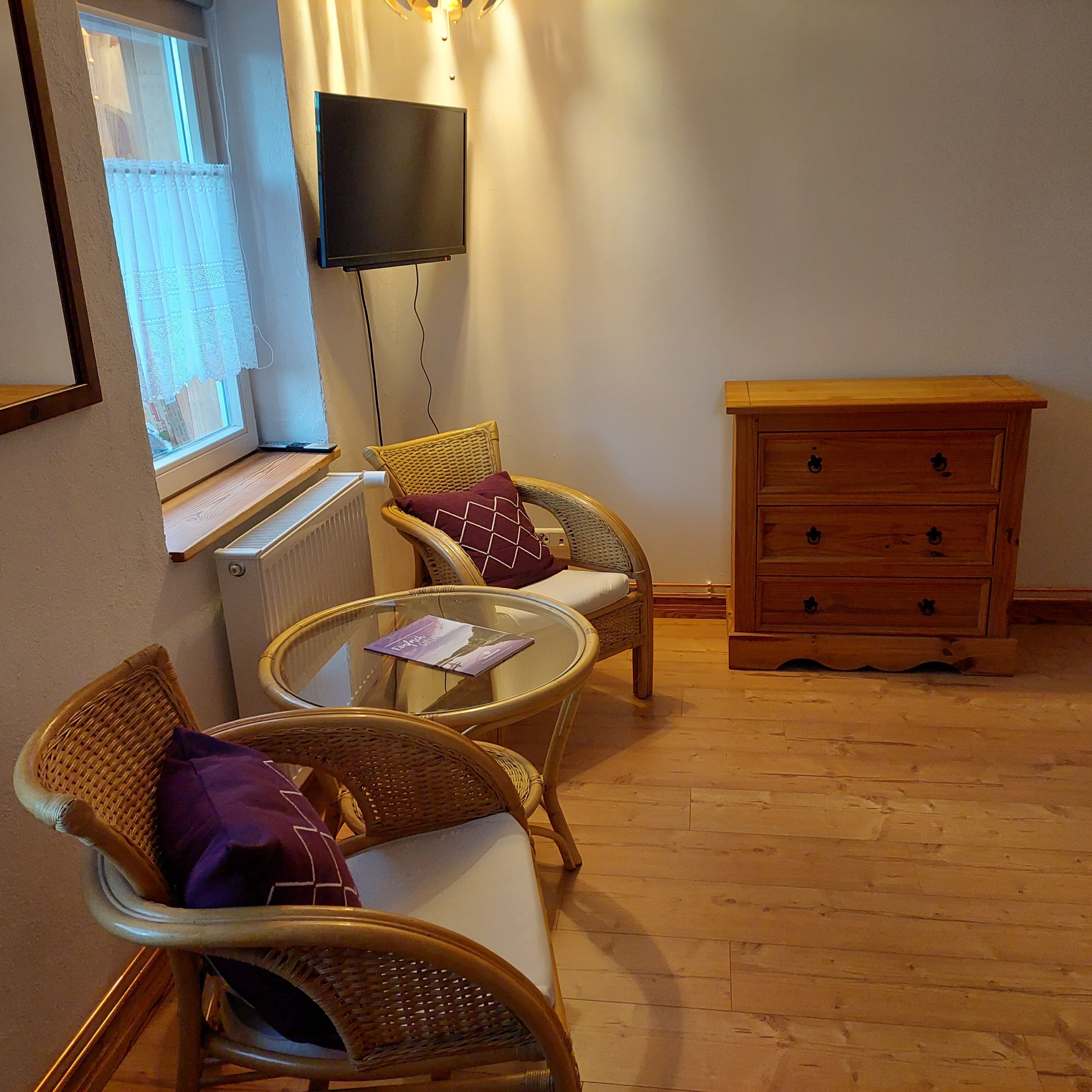Une pièce meublée de meubles en osier, avec une petite table en verre, une commode en bois et un téléviseur fixé au mur.